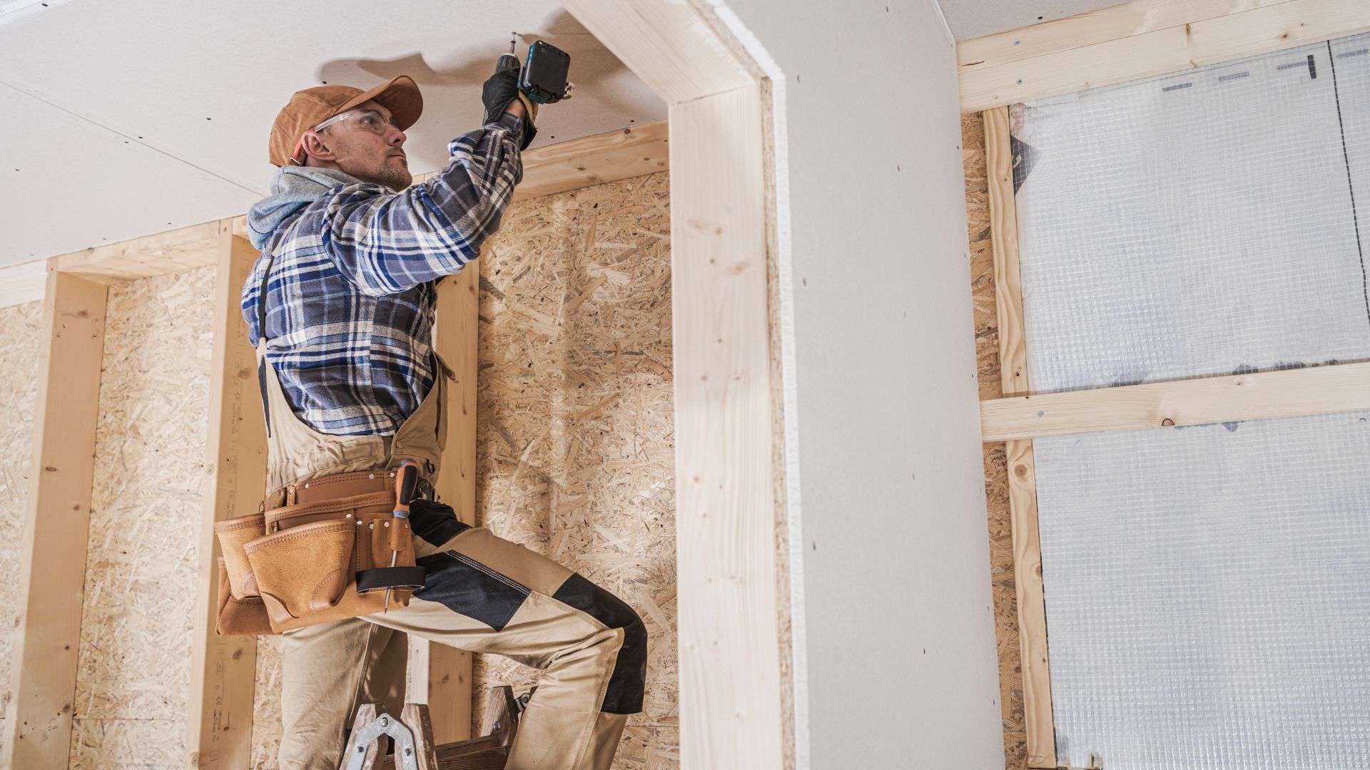 A construction worker in a plaid shirt and cap stands on a ladder, using a tool to work on the ceiling of a wooden frame structure. They have a tool belt around their waist, and the space is partially covered in drywall.