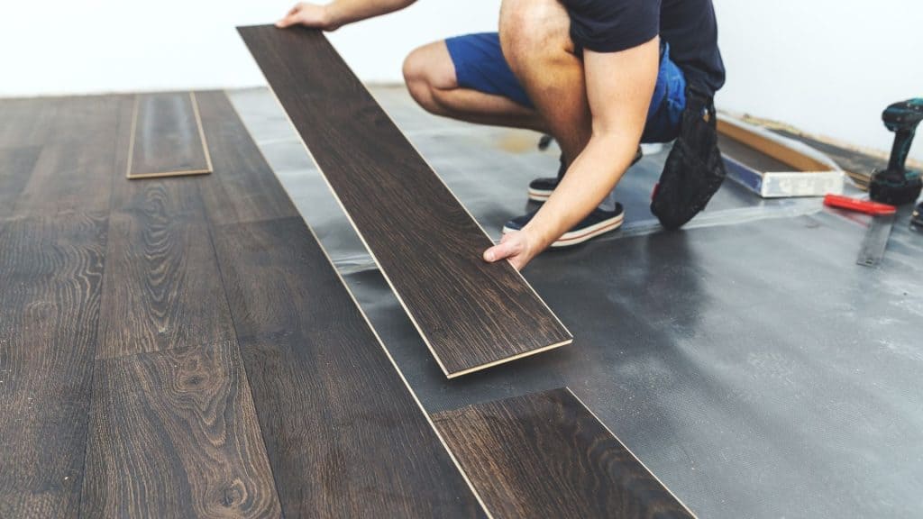 A person kneeling on the floor installing dark wood laminate flooring. Tools and additional planks are visible nearby, indicating a home improvement project in progress. The person is wearing a dark shirt and blue shorts.