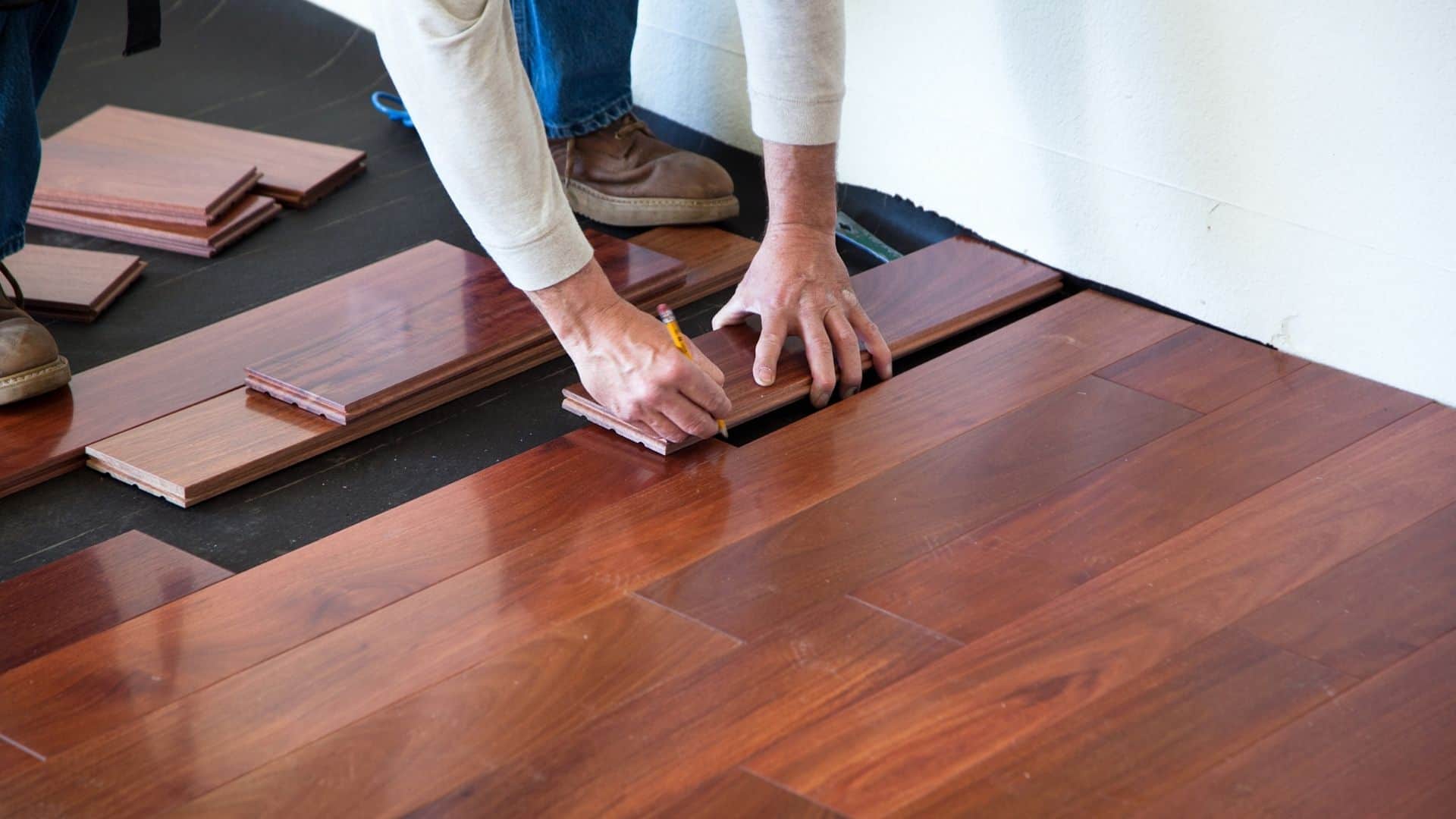 A person installing dark wood laminate flooring, using a measuring tool to align planks. Several wood planks are laid on a subfloor, with hands adjusting their position. The person is wearing jeans and brown shoes.
