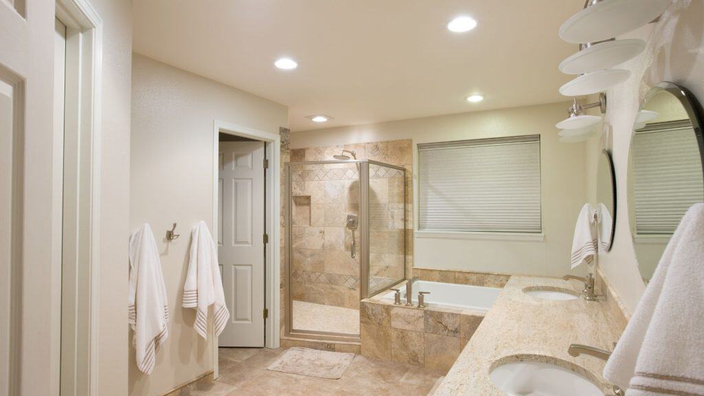 A modern bathroom with beige tiles features a glass-enclosed shower and a bathtub. There are double sinks with a large mirror above, and white towels hang on the walls. Recessed lighting illuminates the space.