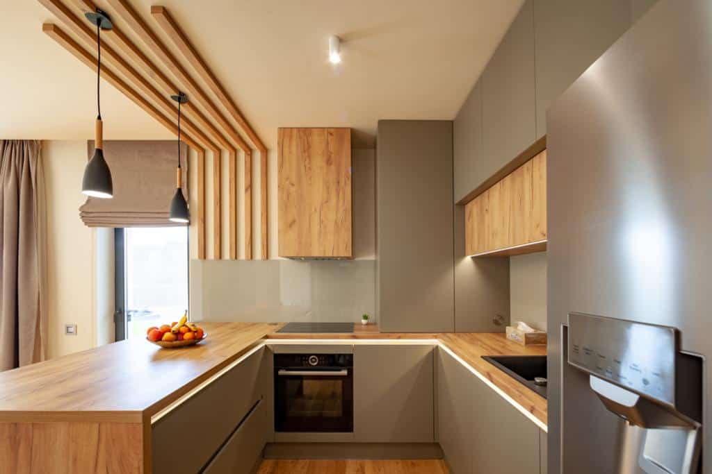 A modern kitchen remodeled by a skilled general contractor features wooden cabinets, a built-in oven, and a black sink. A fruit bowl on the countertop adds color. Pendant lights hang from the ceiling, while natural light flows through a large window framed with a brown curtain.