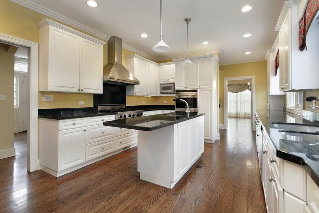 A modern kitchen showcases white cabinetry, black countertops, and stainless steel appliances, ideal for any kitchen remodeling project. It features a central island with a sink and pendant lights above. Hardwood floors extend throughout, while a large window invites natural light in.