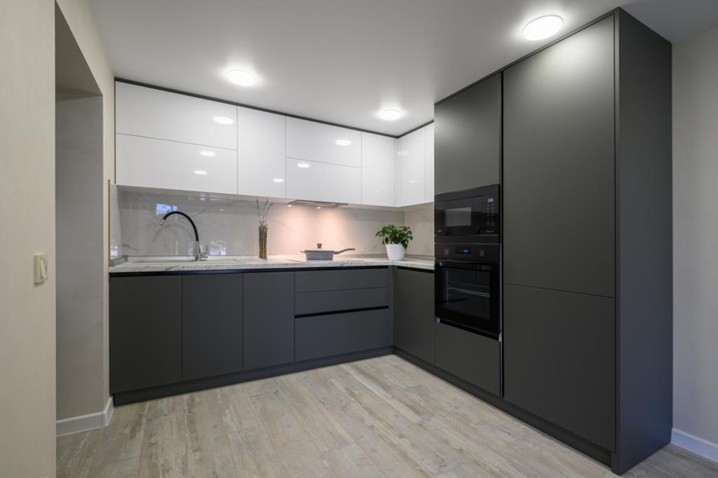 A modern kitchen with dark gray lower cabinets and white upper cabinets. It features a built-in oven and microwave, a sleek countertop with a sink, and a potted plant—perfectly crafted by a general contractor. The light wood floor and recessed ceiling lighting complete the look.