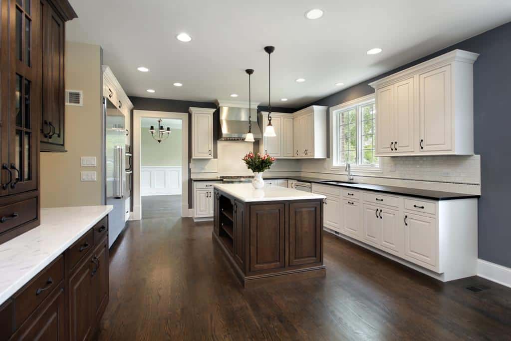 A modern kitchen with dark hardwood floors and gray walls showcases expert kitchen remodeling. It features white cabinets, black countertops, and a central island with rich brown wood. Pendant lights illuminate the space, while a vase with flowers adorns the island. A doorway leads to another room.