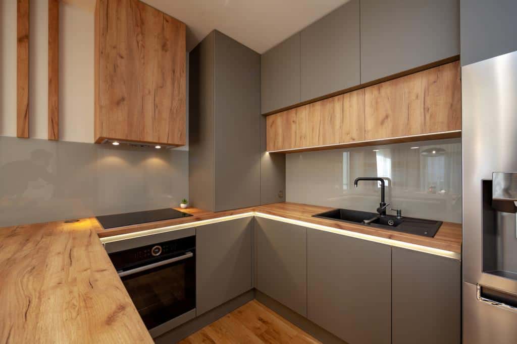 A modern kitchen with wooden countertops and gray cabinets, crafted with the expertise of general contracting. It features a built-in electric stovetop, black sink, and stainless steel refrigerator. The glass backsplash complements the warm lighting for a welcoming space.