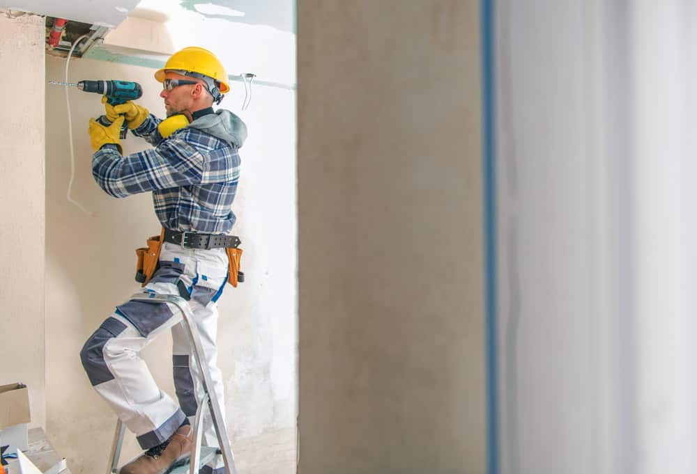 A construction worker wearing a yellow hard hat, safety goggles, and gloves stands on a ladder using a power drill on a wall. He is dressed in a checkered shirt and white work pants with a tool belt around his waist.