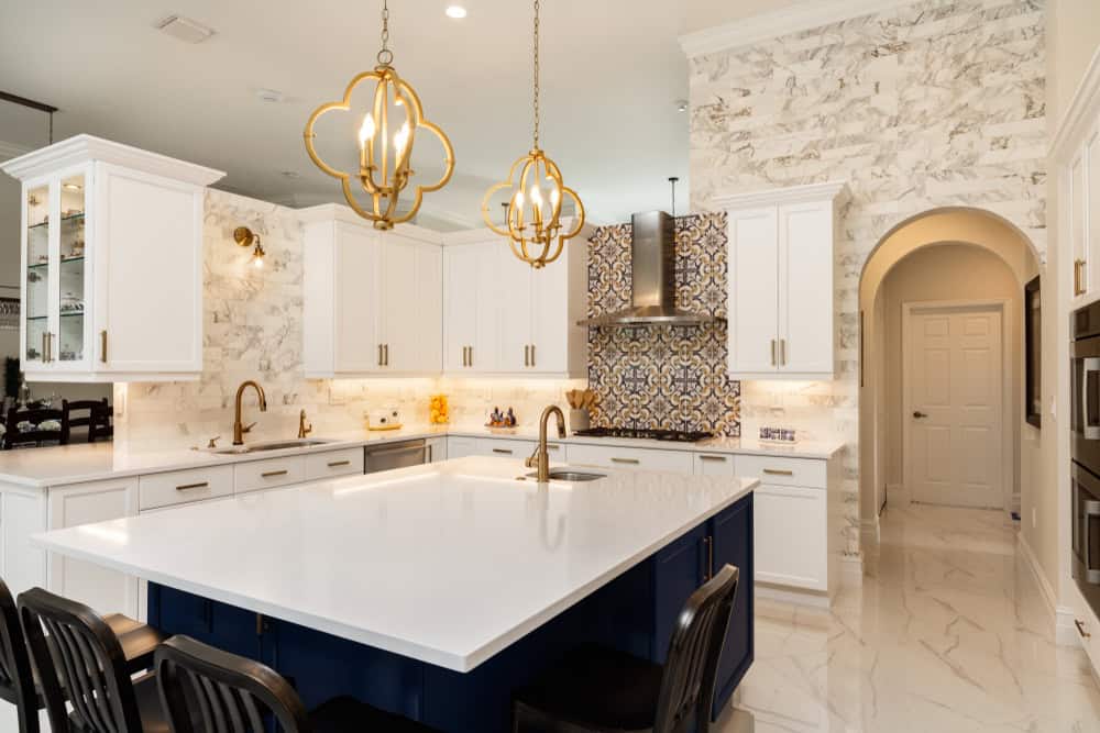 Modern kitchen with white cabinets, marble backsplash, and a blue island featuring a white countertop. Gold pendant lights hang above the island, while patterned tiles accent the stove area. The floor is white with a marble finish.