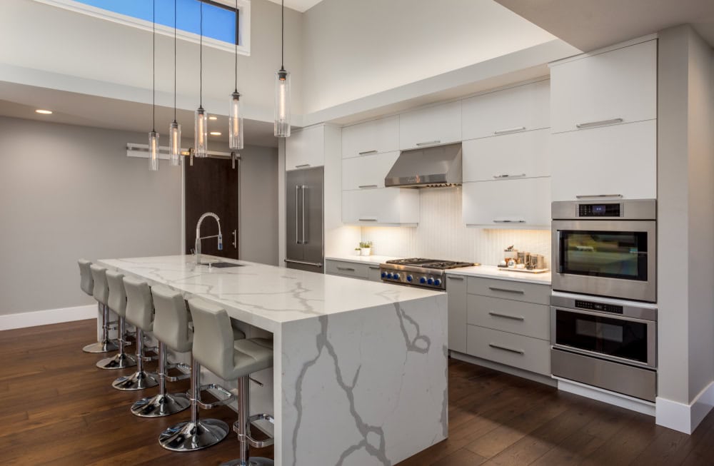 Modern kitchen with white marble island, gray bar stools, and stainless steel appliances. Pendant lights hang above the island. Cabinets are white with sleek handles, and there are wooden floors. High ceiling with a window adds natural light.