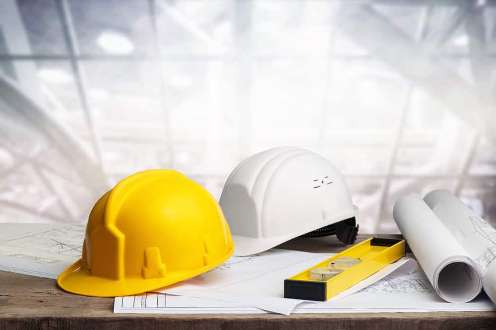 Two hard hats, one yellow and one white, are placed on a wooden table with architectural blueprints and a yellow level tool. The background features a blurred industrial setting with large windows.