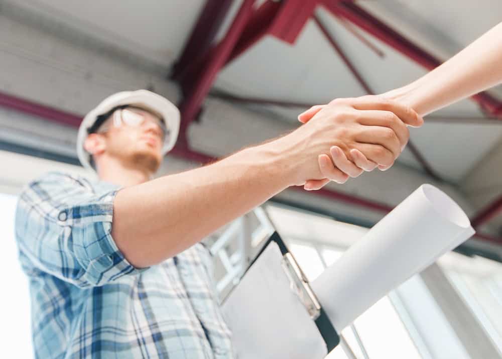 A person in a plaid shirt and construction helmet shakes hands with another person. They hold rolled-up plans under their arm. The background is a building interior with metal beams and natural light.