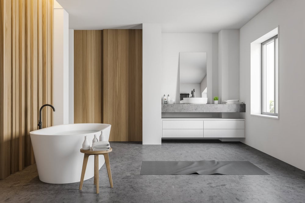 Minimalist bathroom with a freestanding white bathtub, a wooden stool, and a gray mat on the floor. Wooden panel accent wall and a large mirror above a modern white vanity. Large window provides natural light.