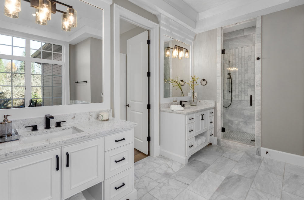 A modern bathroom with gray walls features two white vanities with marble countertops, large mirrors, and black fixtures. A glass-enclosed shower with gray tile and recessed lighting is visible, and there's a window with divided panes.