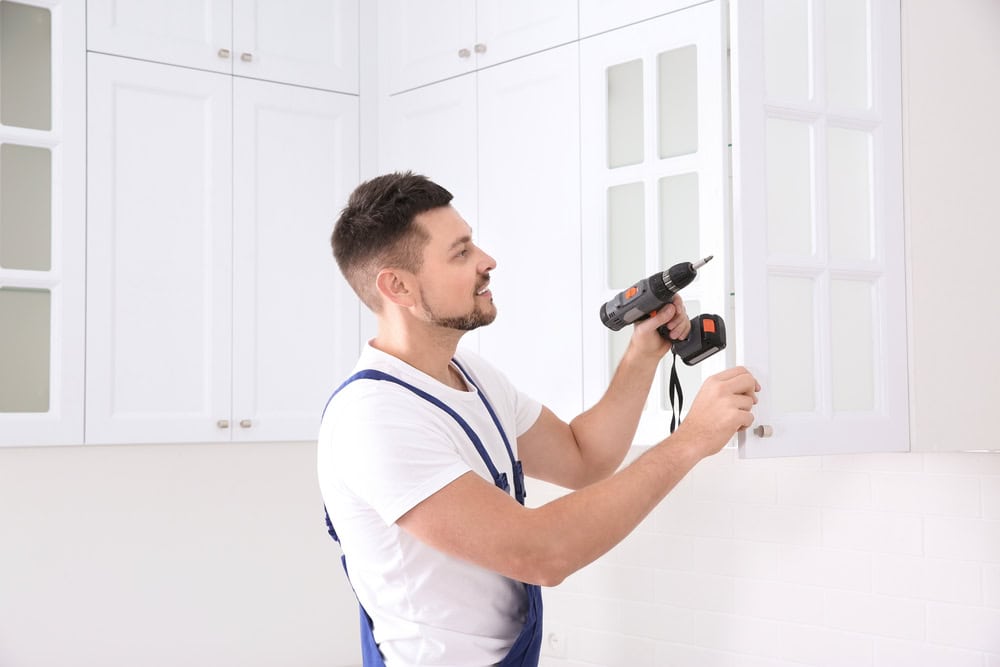A man is wearing a white shirt and blue overalls, using a power drill to install or adjust a white kitchen cabinet. The background shows other white cabinets in a bright, modern kitchen setting.