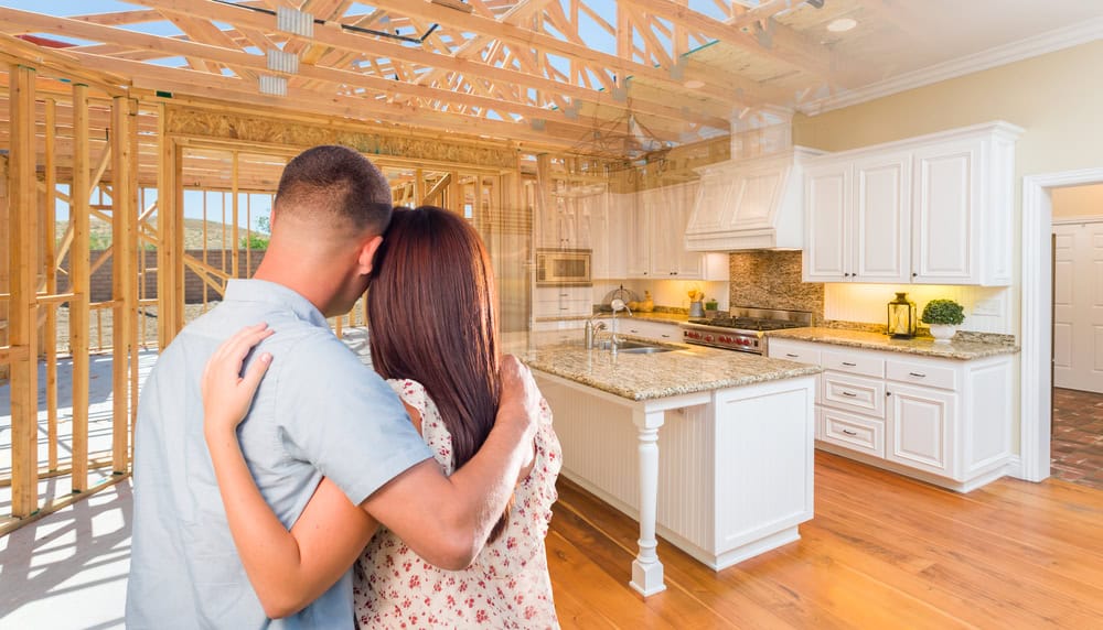 A couple stands with their backs to the viewer, embracing as they gaze at a home under construction. The image blends the unfinished wooden framework with a completed kitchen featuring white cabinets and granite countertops.