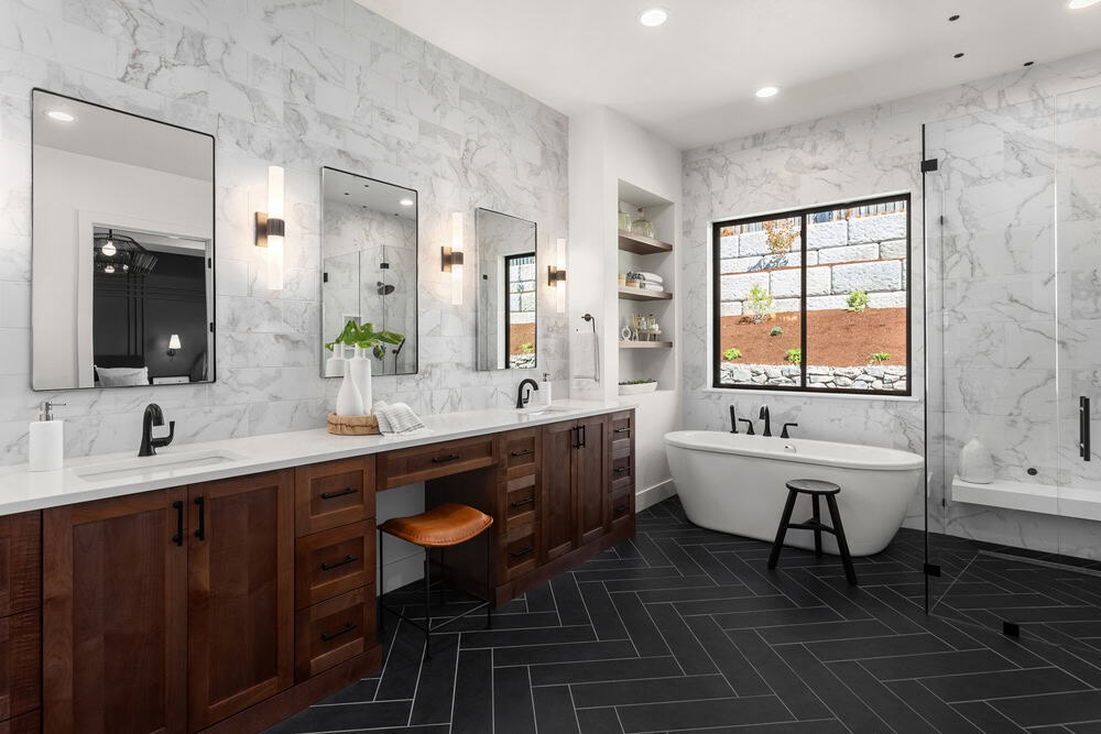 A modern bathroom with dark herringbone tile flooring, marble-patterned walls, and wooden cabinets. It features three mirrors, a freestanding white bathtub, and a walk-in shower. A large window provides natural light.