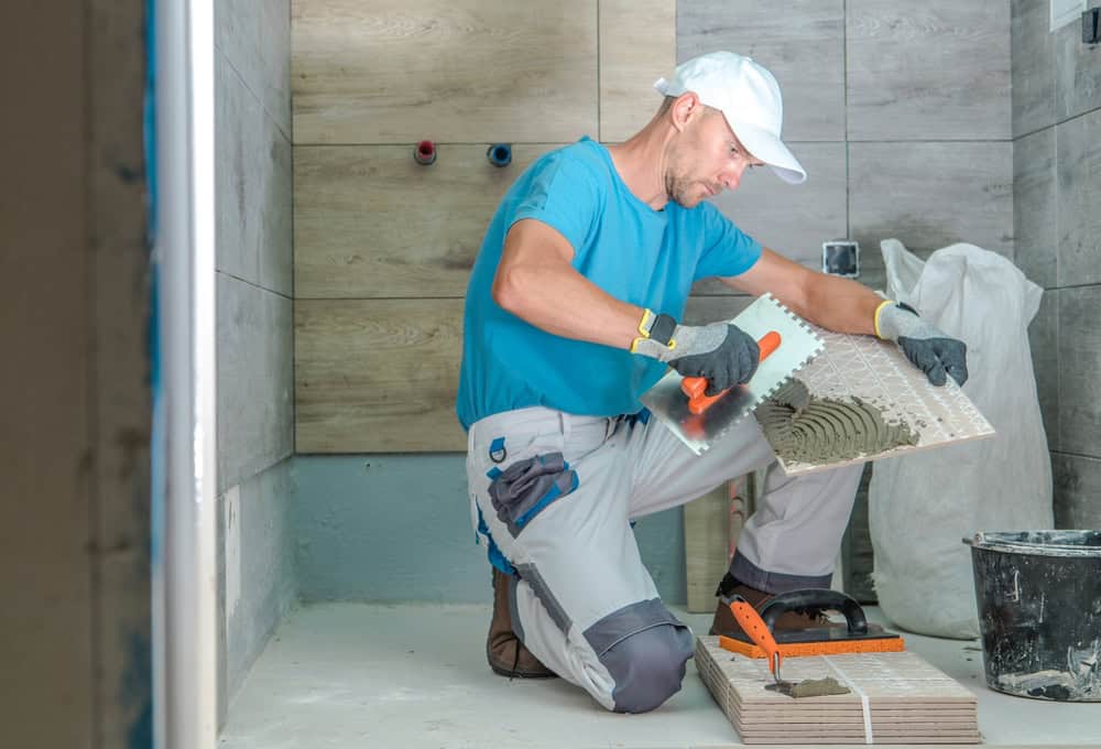 A man wearing a blue shirt and white cap is installing tiles in a bathroom. He is kneeling on the floor and applying adhesive to a large tile with a trowel. The room is under construction with unfinished walls.