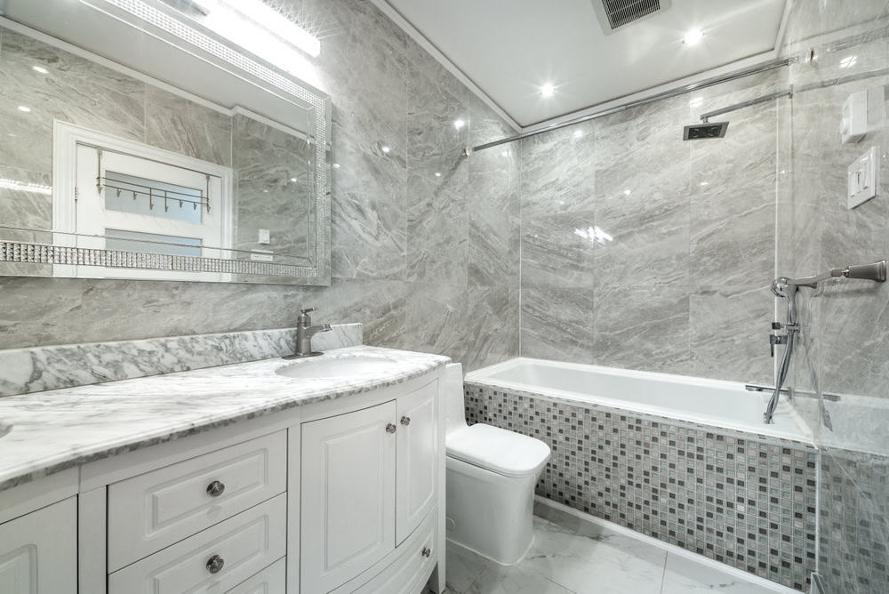 A modern bathroom with marble walls and flooring. It features a white vanity with a marble countertop, a large mirror, a bathtub with mosaic tile detailing, and recessed lighting.