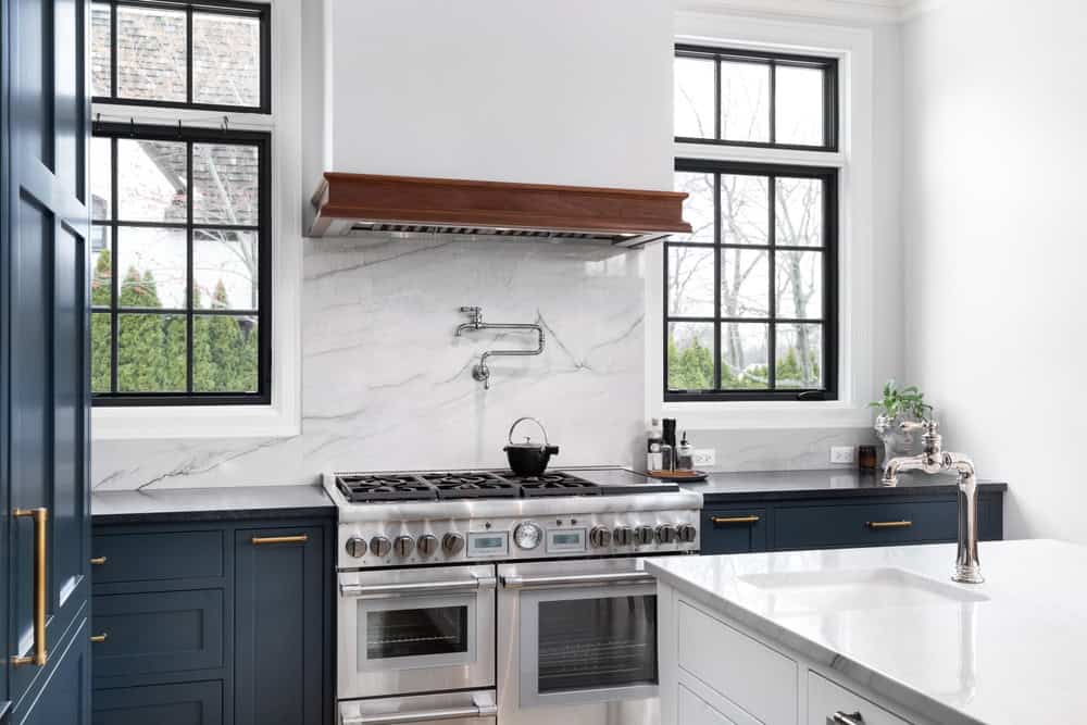 A modern kitchen with navy blue cabinets, a large stainless steel stove, and a marble backsplash. A pot sits on the stove under a pot filler. Windows with black frames provide natural light. A silver faucet is visible on the white island countertop.