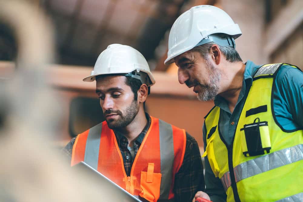 Two construction workers in hard hats and safety vests are reviewing documents on a clipboard. One worker has a gray beard and wears a green vest; the other is bearded with an orange vest. They appear focused and engaged in a discussion.