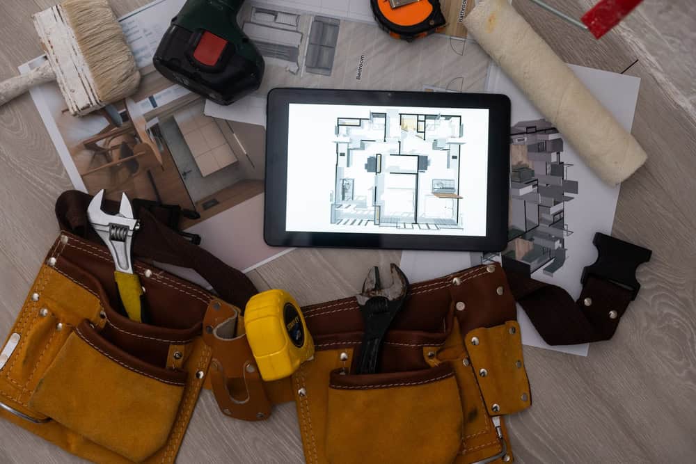 A tablet displaying blueprints lies on a wooden surface surrounded by tools like a wrench, pliers, screwdriver, measuring tape, and a tool belt. Paintbrushes and a roller are also nearby, suggesting home renovation or construction work.