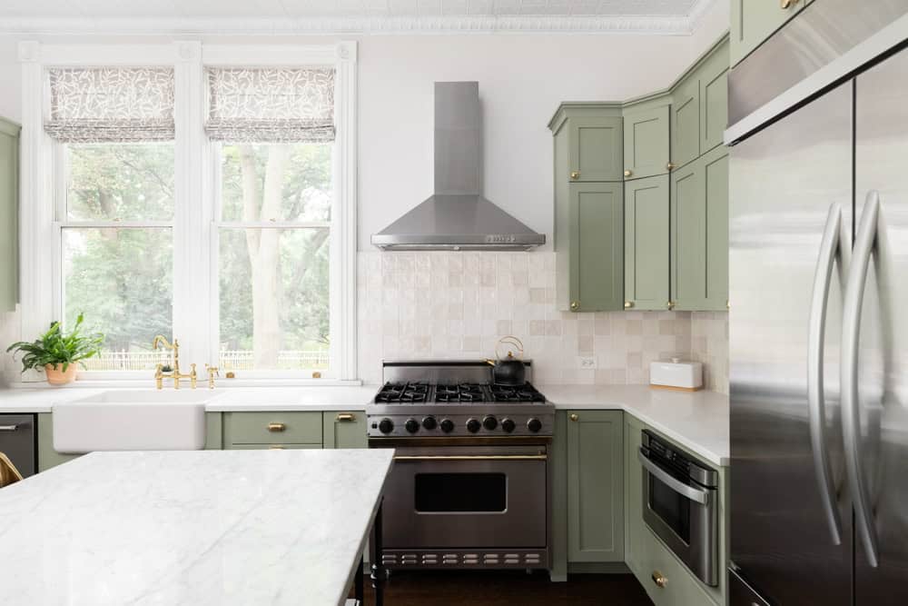 A modern kitchen featuring green cabinetry, a silver range hood, and stainless steel appliances. Light flows through large windows with patterned blinds, and a plant sits next to a farmhouse sink with a gold faucet. A marble countertop island is in view.