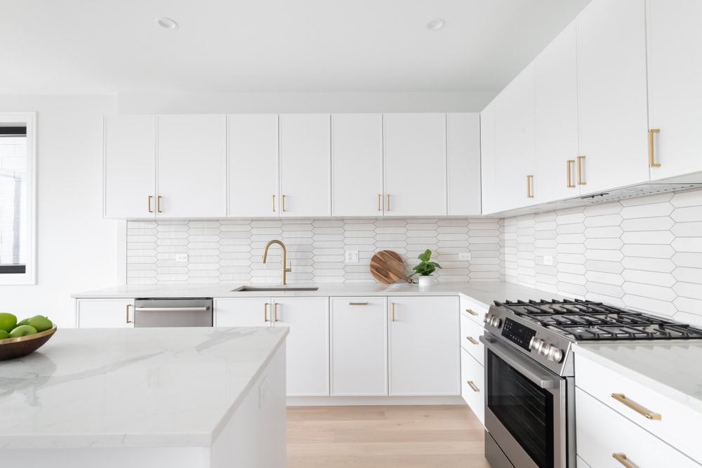 A modern kitchen with white cabinets, a white hexagonal tile backsplash, a stainless steel stove, and a dishwasher. A wooden bowl with greens is on the countertop, alongside a silver faucet and a bowl of green apples.