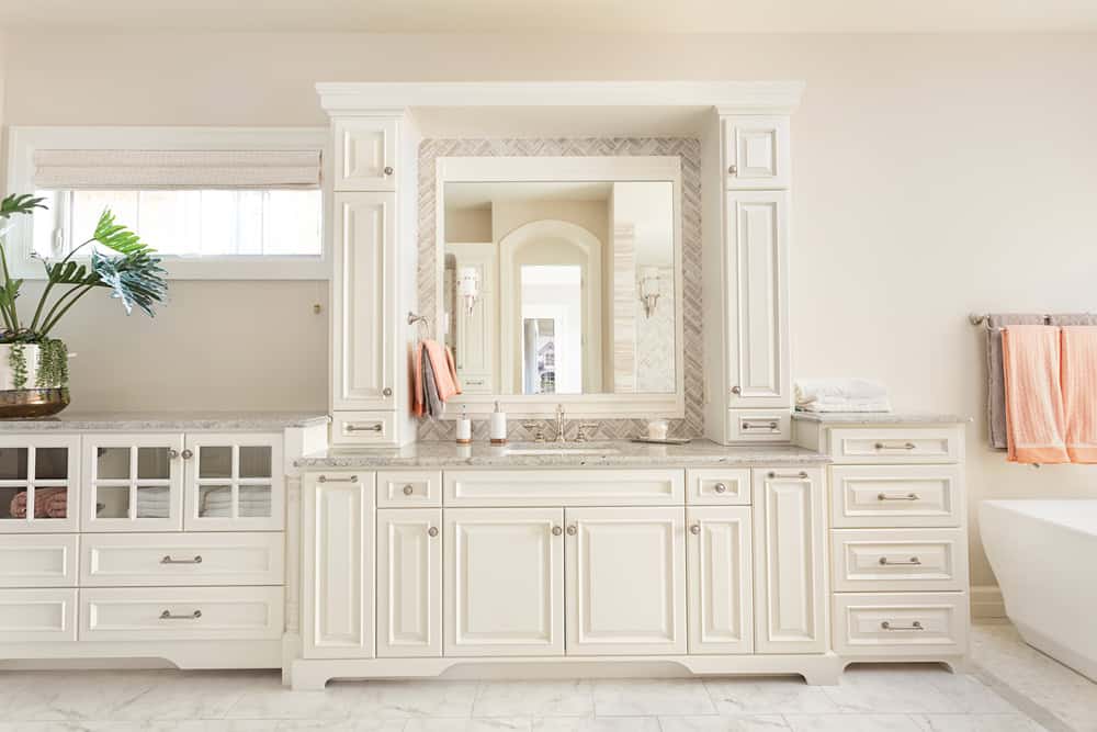 A bright, elegant bathroom features a large white vanity with a marble countertop and a mirror, flanked by tall cabinets. Soft peach towels hang by a white tub. Light tiles and a potted plant complete the serene atmosphere.