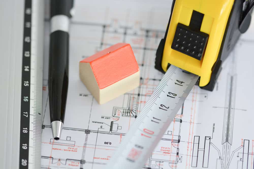 A close-up of a small model house on architectural blueprints. A pen and a yellow measuring tape with an extended metallic strip are nearby, suggesting construction or design planning.