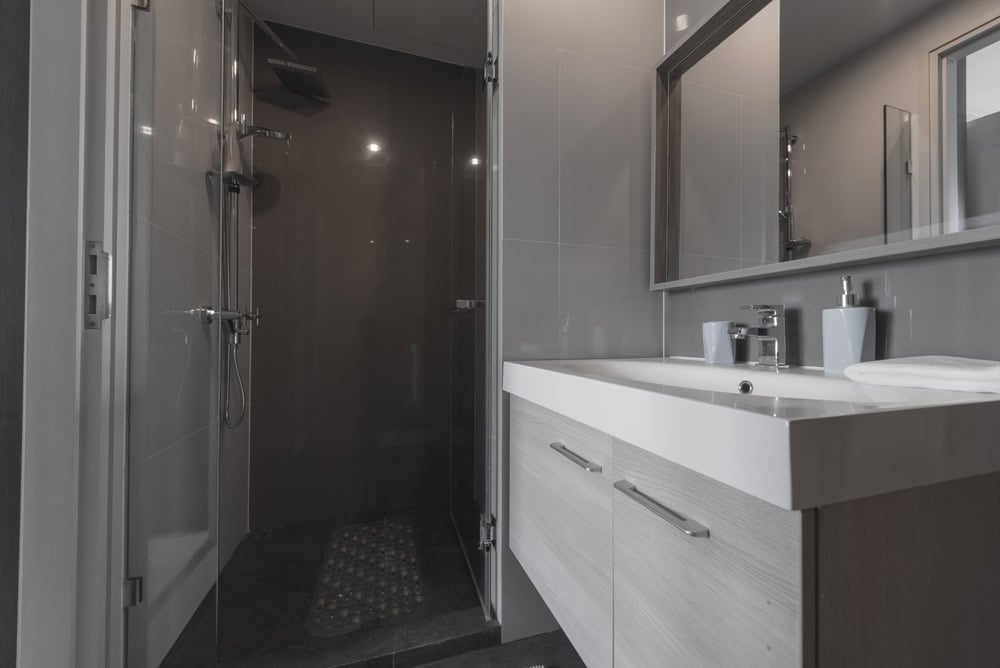 A modern bathroom with a glass-enclosed shower featuring a rain showerhead. The room has gray tiles, a large mirror above a white sink, and wooden cabinets. The countertop holds a soap dispenser and a folded towel.