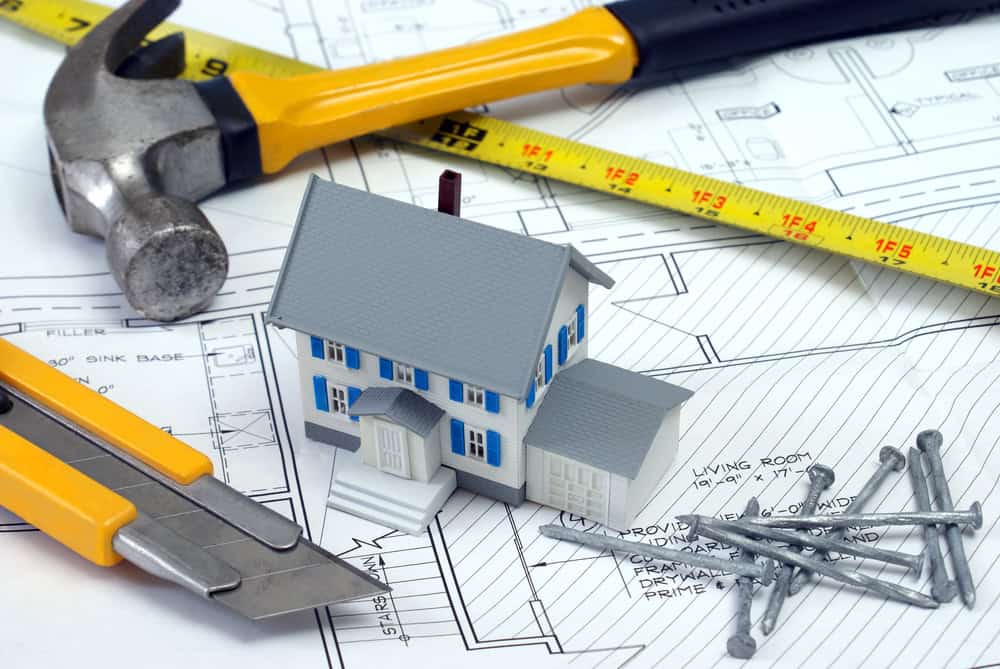 A small model house sits on architectural blueprints, surrounded by a hammer, yellow tape measure, utility knife, and nails. The image represents construction, planning, and home improvement.