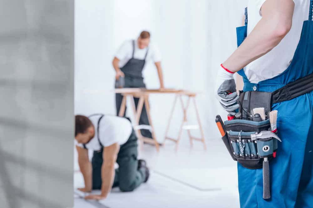 Three workers are in a room under construction. One stands in the foreground with a tool belt, while two others are in the background; one is kneeling, and the other is leaning over a workbench. They all wear blue overalls and white shirts.