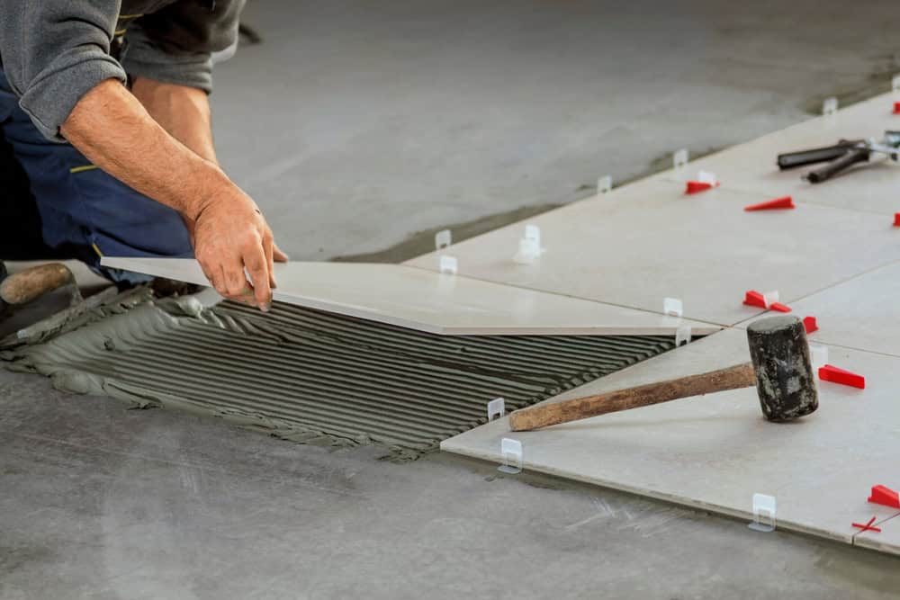 A person is laying large rectangular tiles on a floor using adhesive, with a mallet and red spacers nearby for alignment. The image shows a close-up of the hands placing a tile carefully on the adhesive.