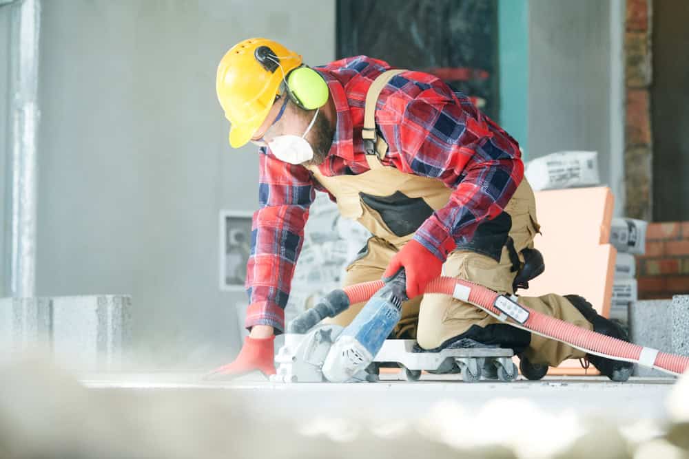 A construction worker wearing a yellow hard hat, ear protection, and a mask is using a power saw on a concrete surface. He's dressed in a red plaid shirt, overalls, and red gloves, surrounded by construction materials in a building site.