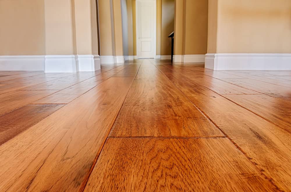Close-up of a polished wooden floor leading to a white door in a hallway. The floors are shiny with warm brown tones, and the walls are a soft beige with white baseboards. The door is centered at the end of the hall.