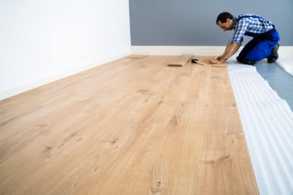 A person kneels on the floor, installing light brown wooden flooring in a room with white and gray walls. They are wearing a blue checkered shirt and blue overalls, working with tools and planks.