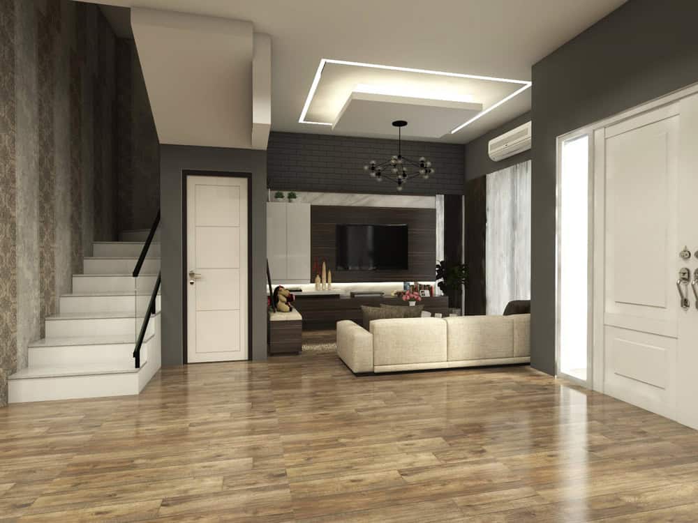 Modern living room with wooden floors, a beige sofa facing a flat-screen TV on a wall unit, and large windows. A staircase with a black railing is on the left. Ceiling features recessed lighting. Walls are a combination of patterned and plain paint.