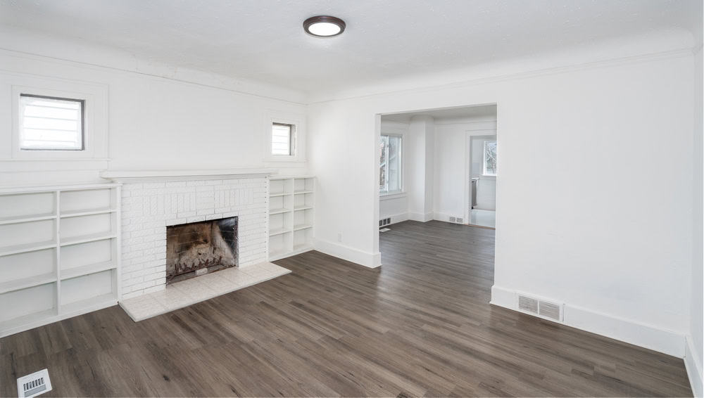 A white living room with a brick fireplace surrounded by empty built-in shelves. The room has light gray wood flooring and a simple circular ceiling light. A doorway leads to an adjoining room with a large window.